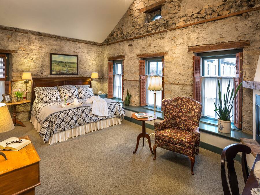 A bedroom with a stone wall, a bed with patterned bedding, a chair, a table with a lamp, and a window with shutters.