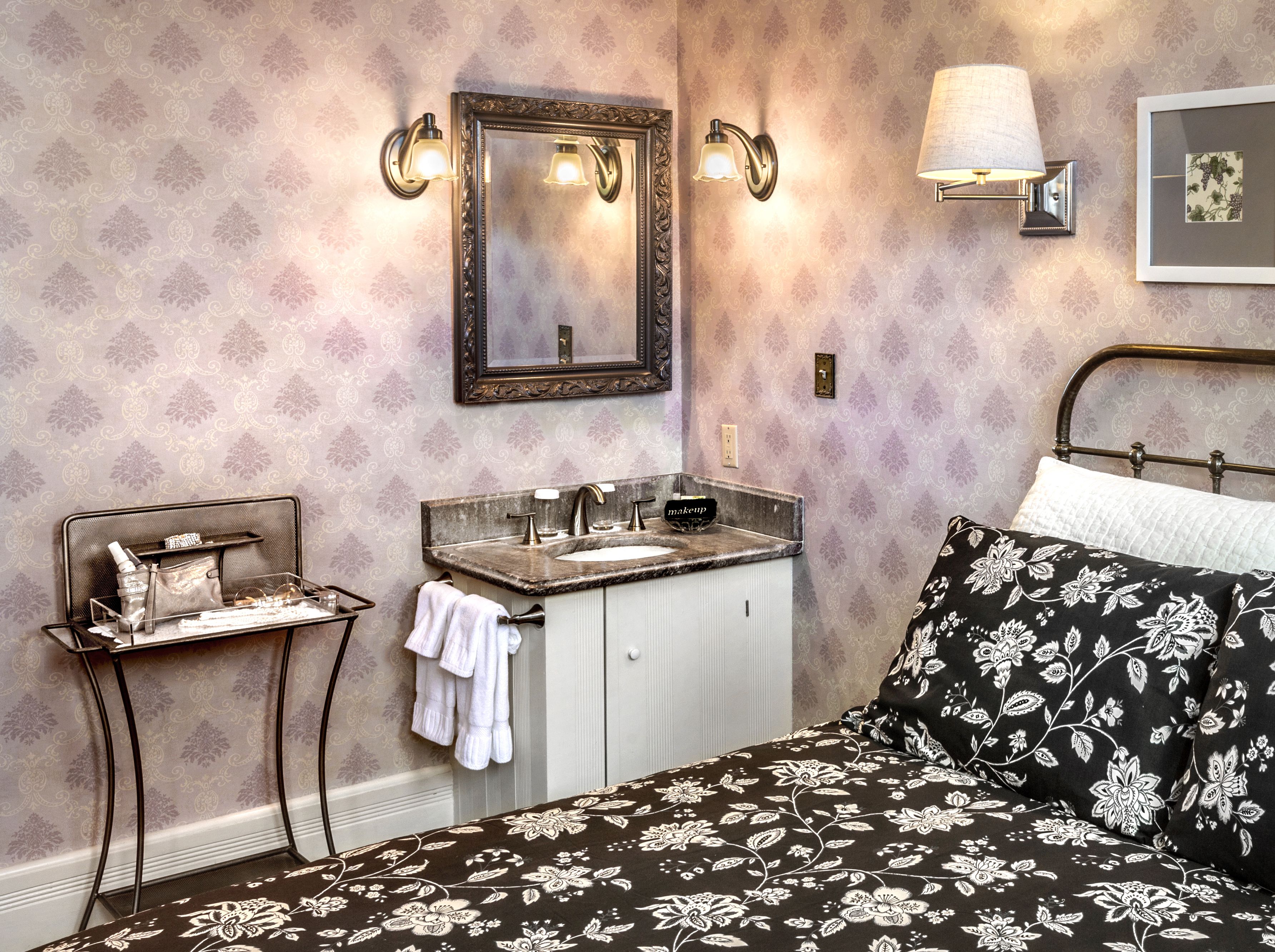 A bathroom with a vanity, sink, and mirror. There is a bed with a floral bedspread and a pillow.