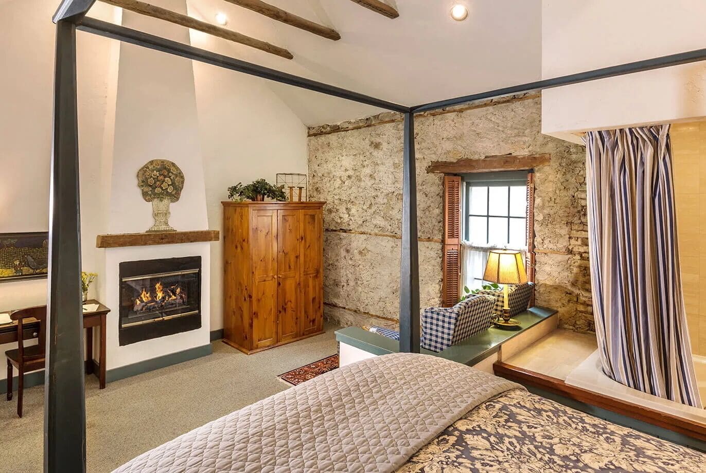 A bedroom with a fireplace, a wooden wardrobe, a bedside table with a lamp, a striped curtain, and a bed with a quilted blanket.