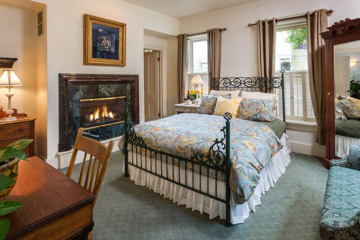 A bedroom with a bed, a fireplace, a nightstand, a chair, and a window with curtains.