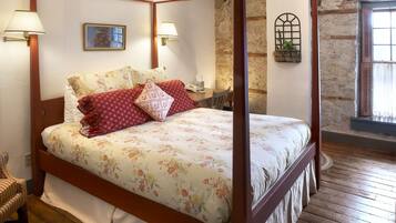 A four-poster bed with floral bedding, a wooden floor, a stone wall, and a window with a curtain.