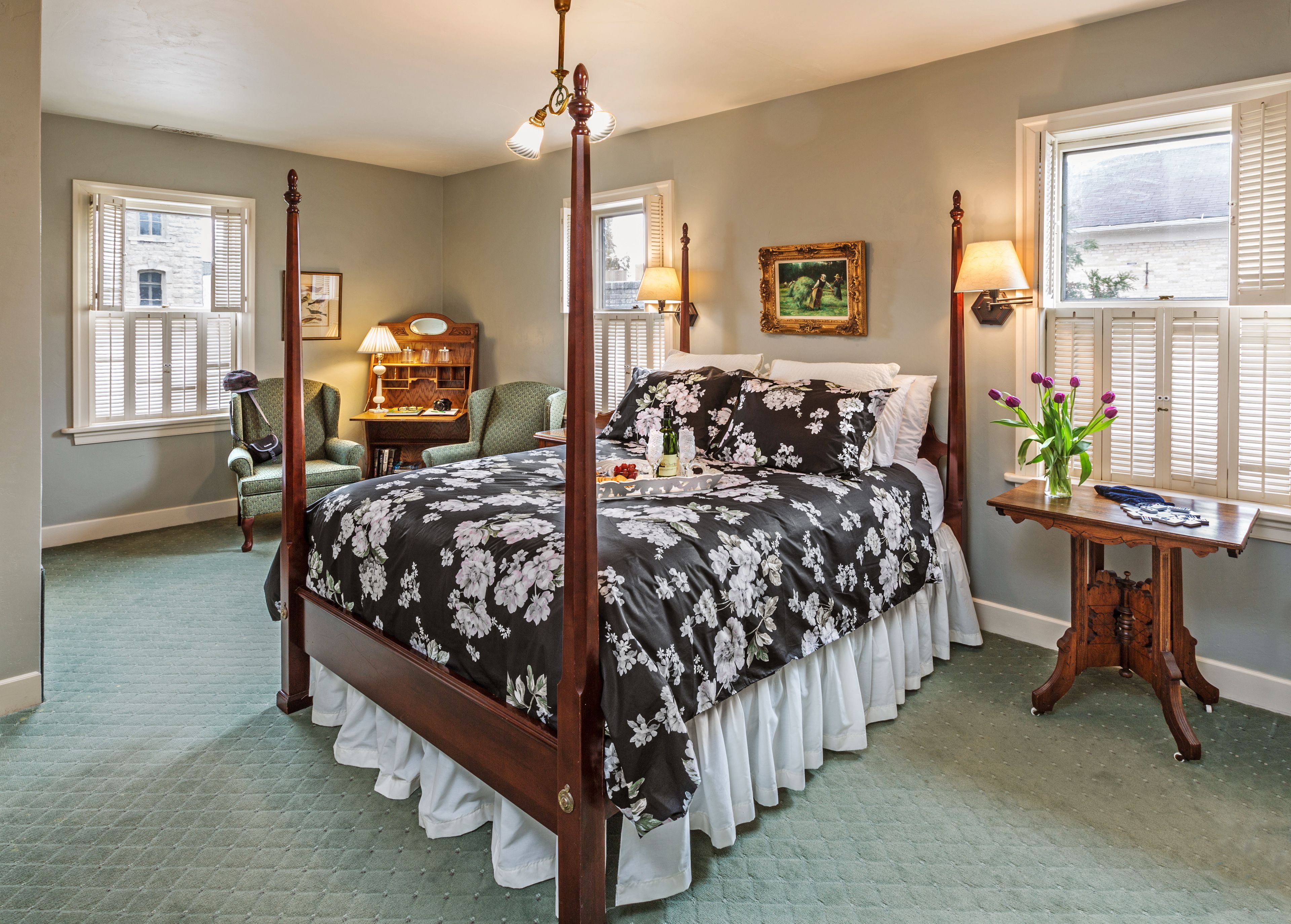 A four-poster bed with a floral bedspread, a wooden nightstand with a vase of flowers, and a desk with a lamp.