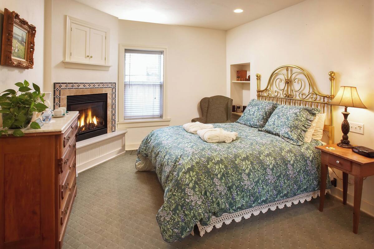 A bedroom with a bed, a dresser, a chair, a bedside table, a lamp, a fireplace, and a window.