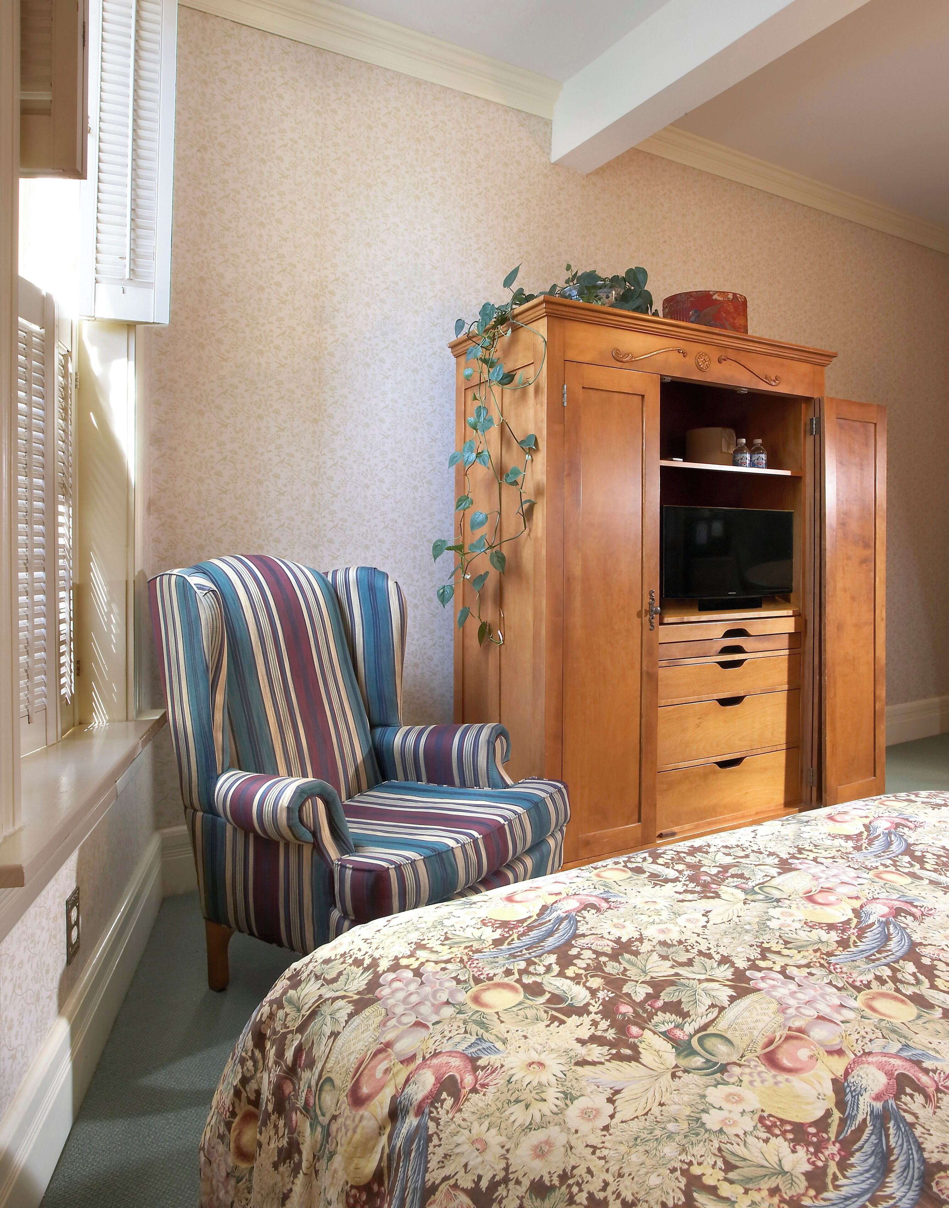 A bedroom with a striped armchair, a wooden wardrobe with a television, and a floral bedspread.