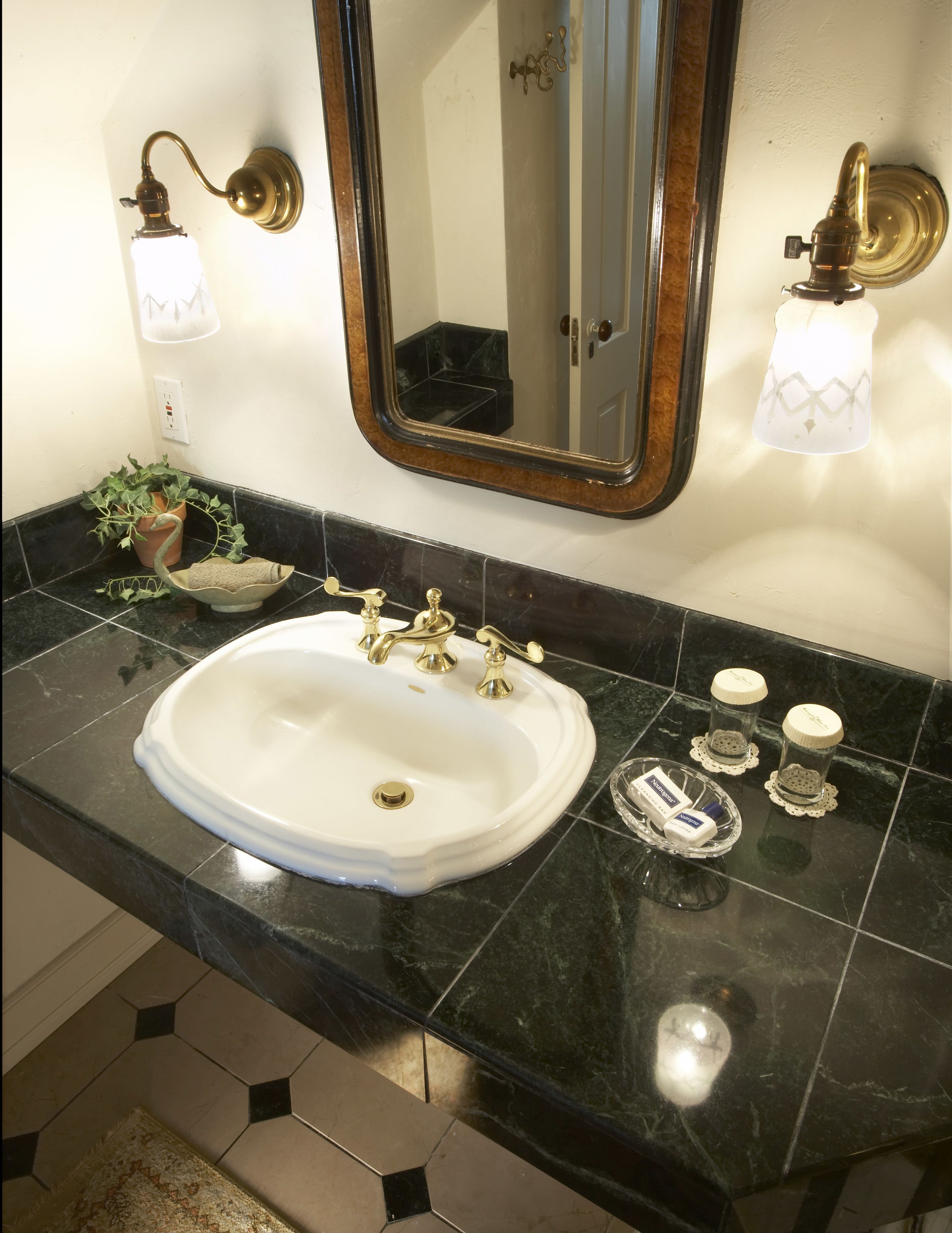 A bathroom with a white sink, gold faucets, and black marble countertops. There are two wall-mounted lights, a mirror, and a potted plant.