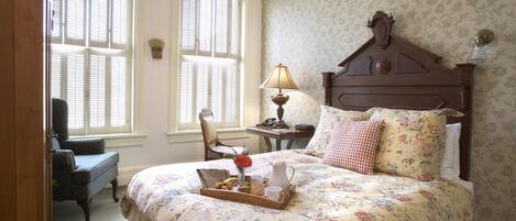 A bedroom with a floral bedspread, a wooden headboard, a nightstand with a lamp, and a chair.