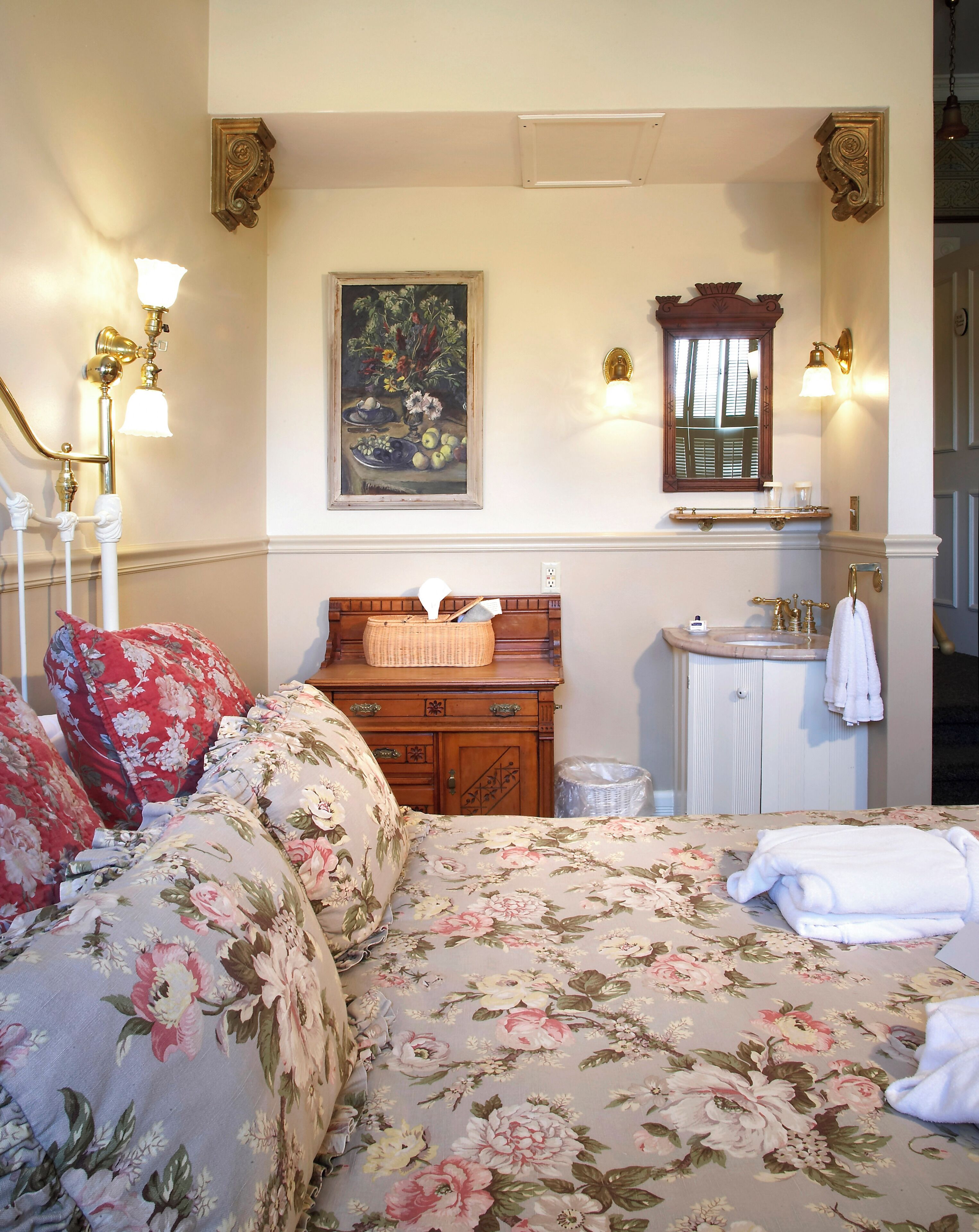 Un dormitorio con cubrecama floral, cómoda de madera, tocador con espejo y un cuadro en la pared.