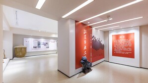 Een modern interieur met een strak, minimalistisch design, met een rode accentmuur in het midden met tekst en een sculptuur, en een vitrine met informatiepanelen.
