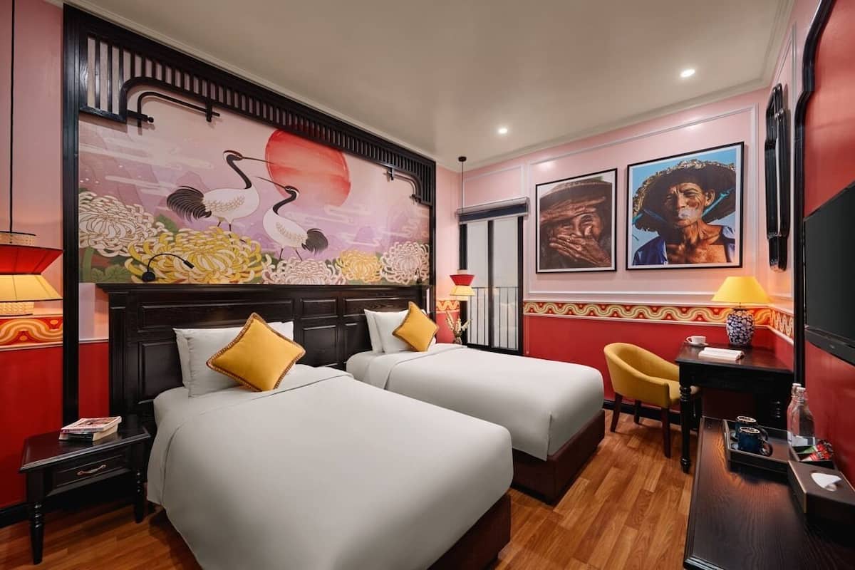 A hotel room with two beds, a wooden floor, a red accent wall, a large painting, and a small table with a lamp.