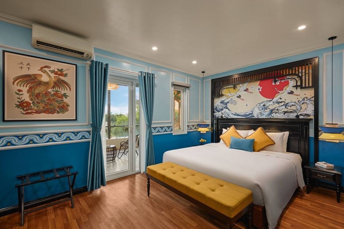 A bedroom with a large bed, a bench, a window with curtains, and artwork on the walls.
