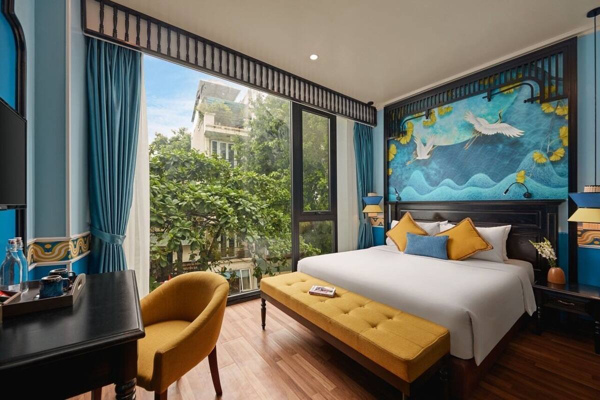A hotel room with a large bed, a bench, a desk, and a view of greenery outside.