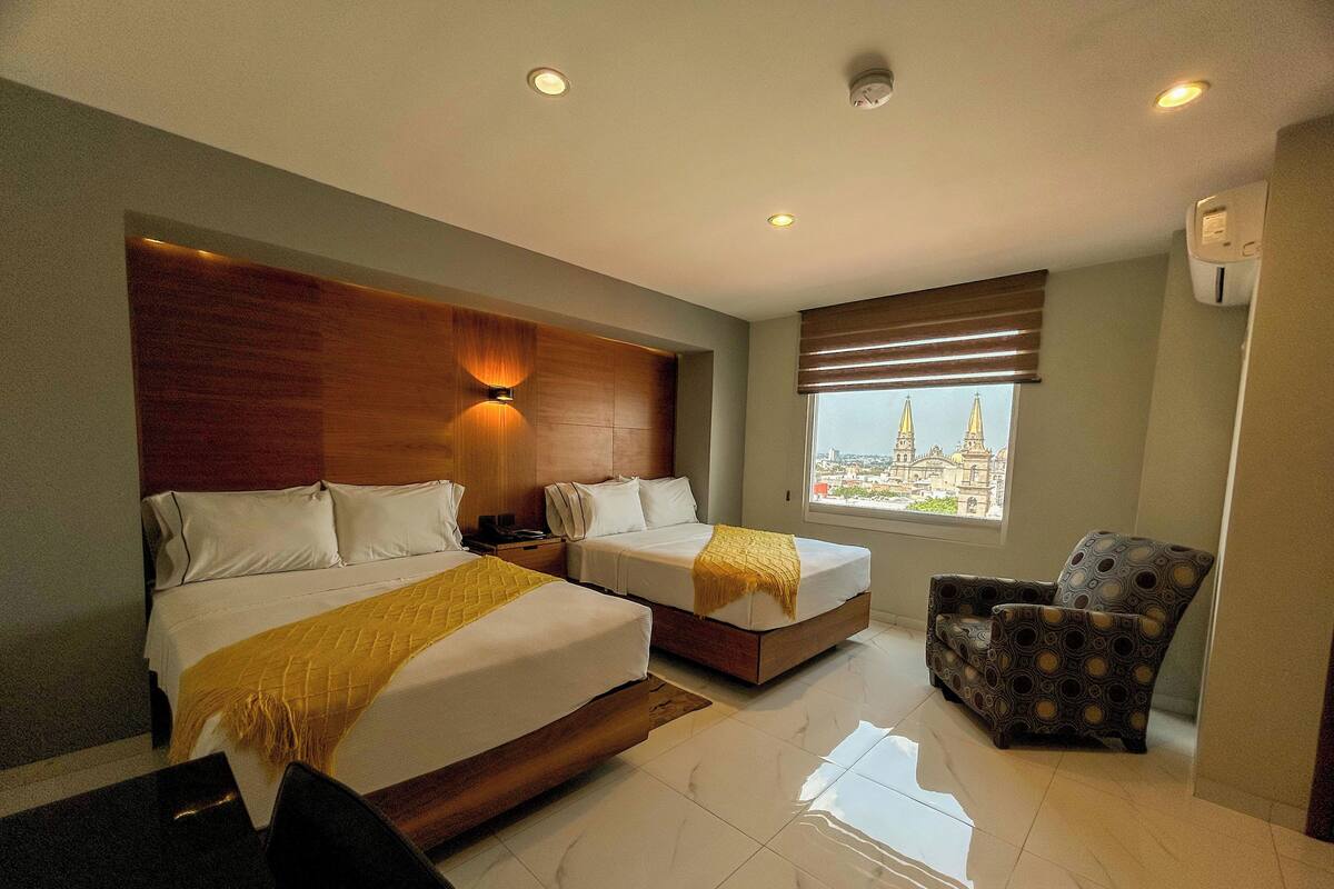Ein Hotelzimmer mit zwei Betten, einem hölzernen Kopfteil, einem großen Fenster mit Blick auf die Stadt, einem gemusterten Sessel und einem kleinen Tisch.