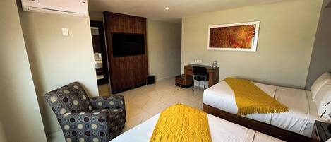 A hotel room with two beds, a desk, a chair, an air conditioning unit, and a television.