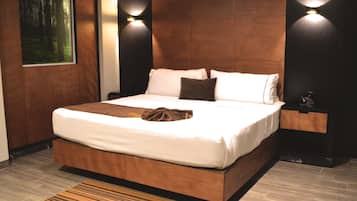 A modern bedroom with a large bed, wooden headboard, two wall-mounted lights, a nightstand, and a view of a forest through a window.