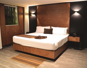 Un dormitorio moderno con una cama grande, cabecera de madera, dos luces fijadas a la pared, una mesita de noche y vistas a un bosque por la ventana.