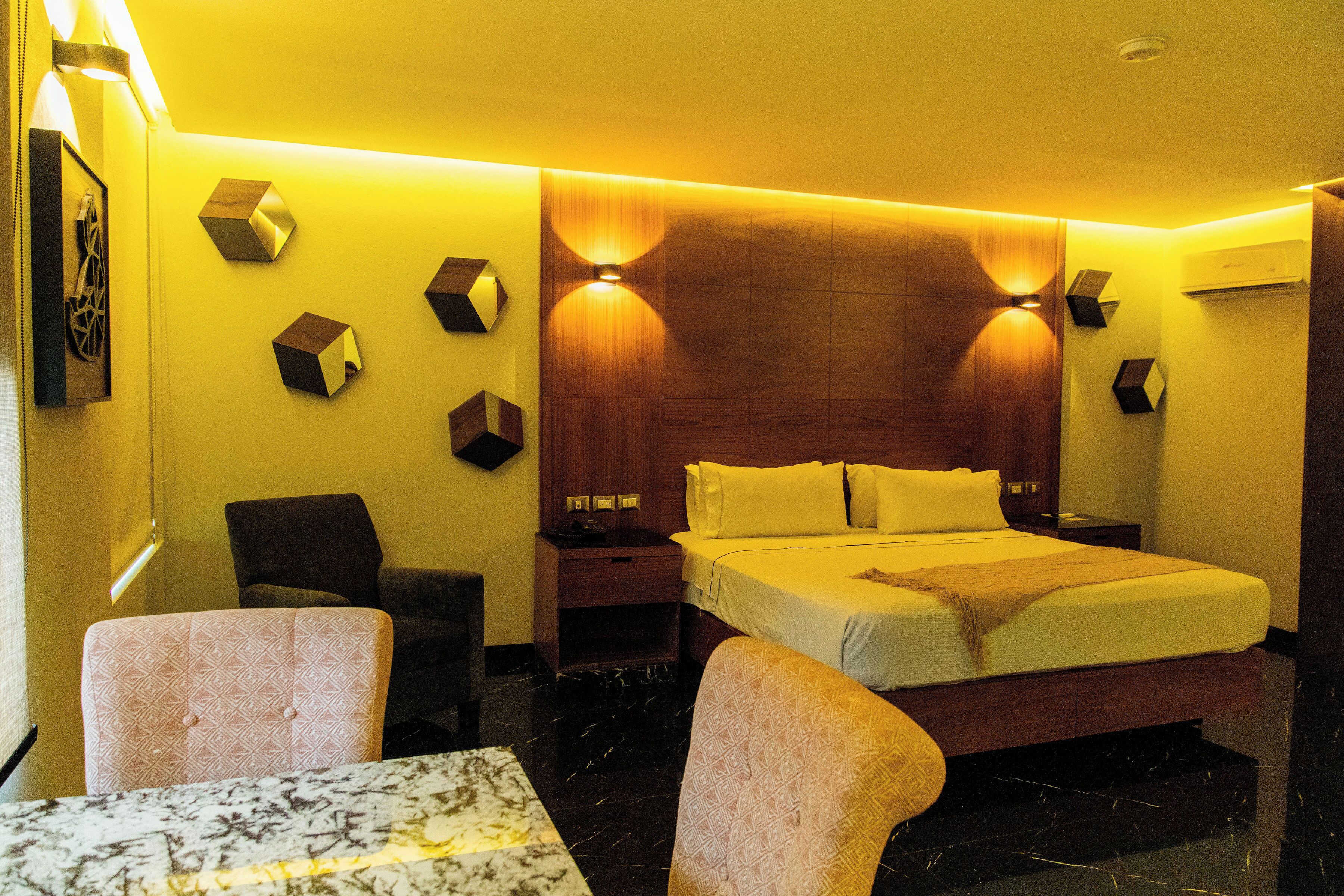 A hotel room with a bed, two chairs, a table, and a wooden headboard with wall-mounted lights.