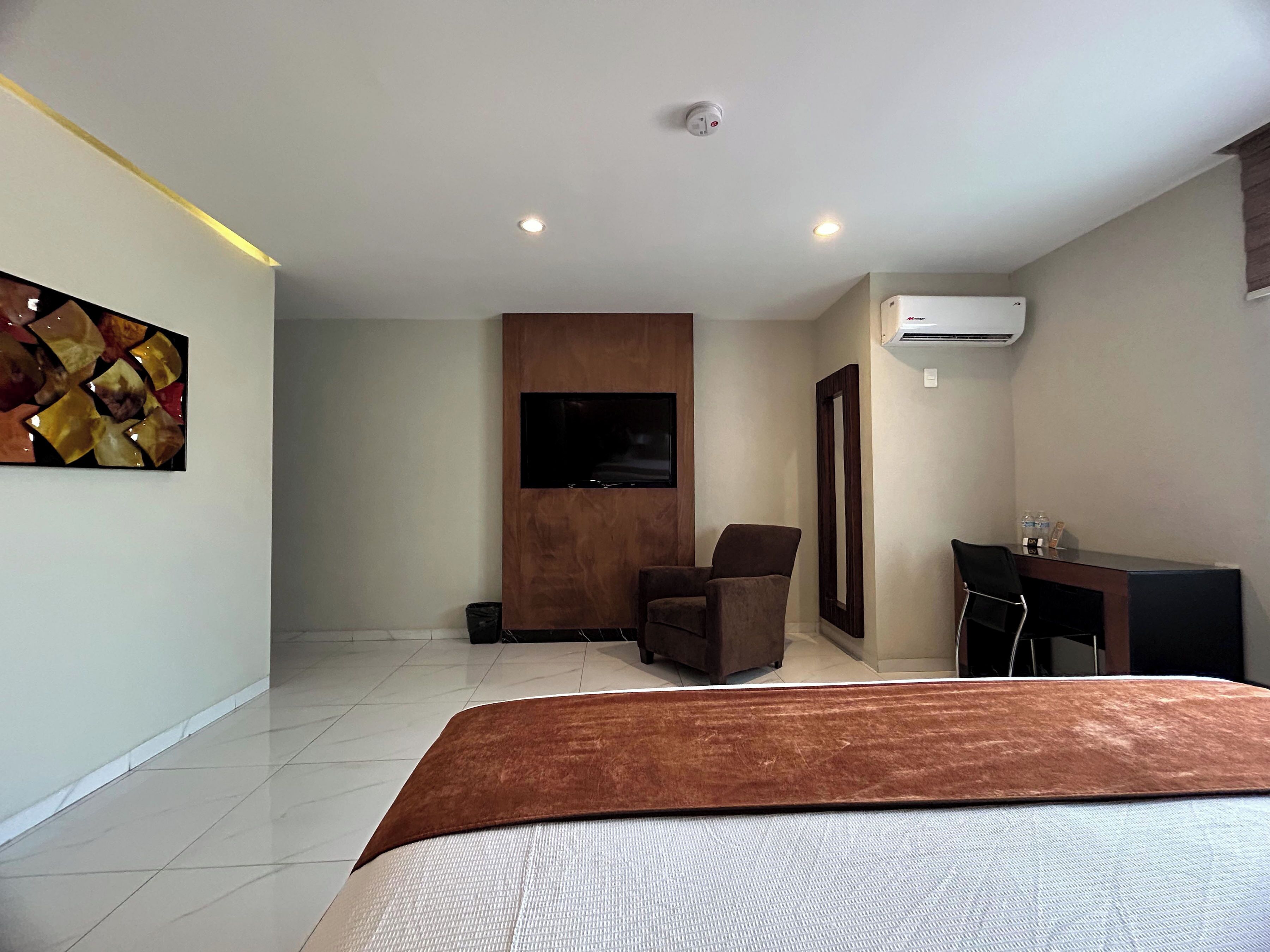 A hotel room with a bed, a desk, a chair, a TV, and an air conditioning unit.