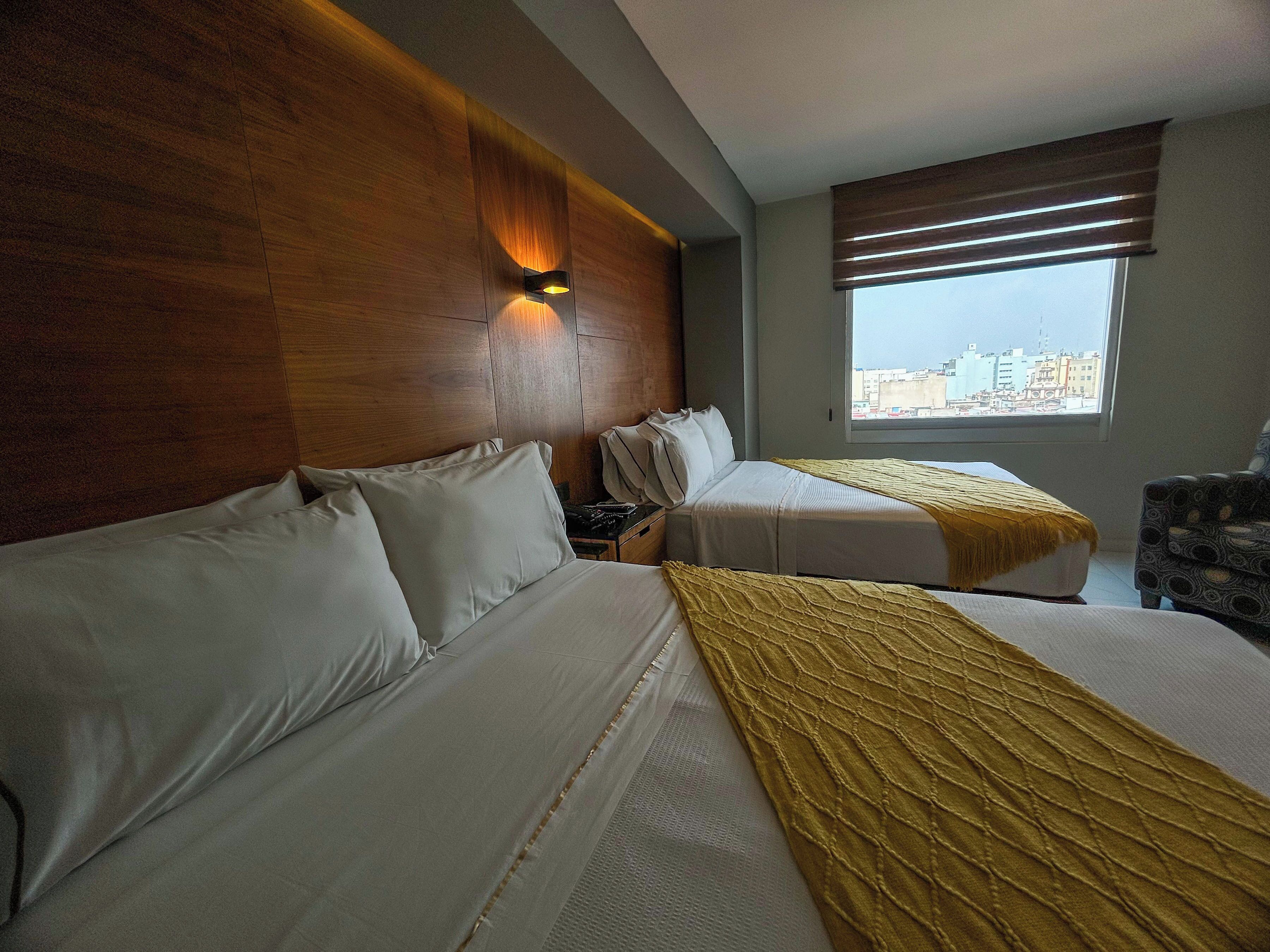 A hotel room with two beds, a wooden headboard, a nightstand, a lamp, a window with blinds, and a view of the cityscape.