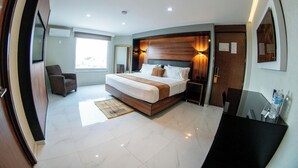 A modern hotel room with a large bed, a sitting area with a chair, and a desk with a water bottle.