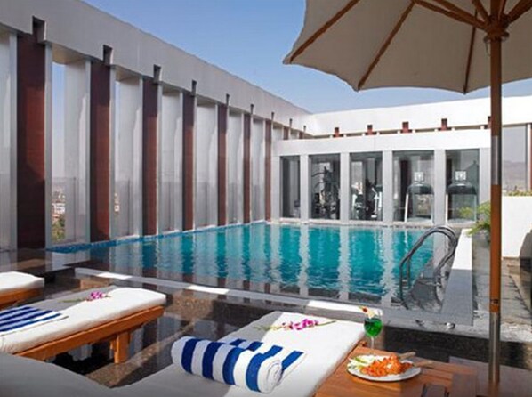 A modern indoor pool area with a clear view of the pool, lounge chairs, and a table with a drink. - Royal Orchid Central Grazia (Navi Mumbai)