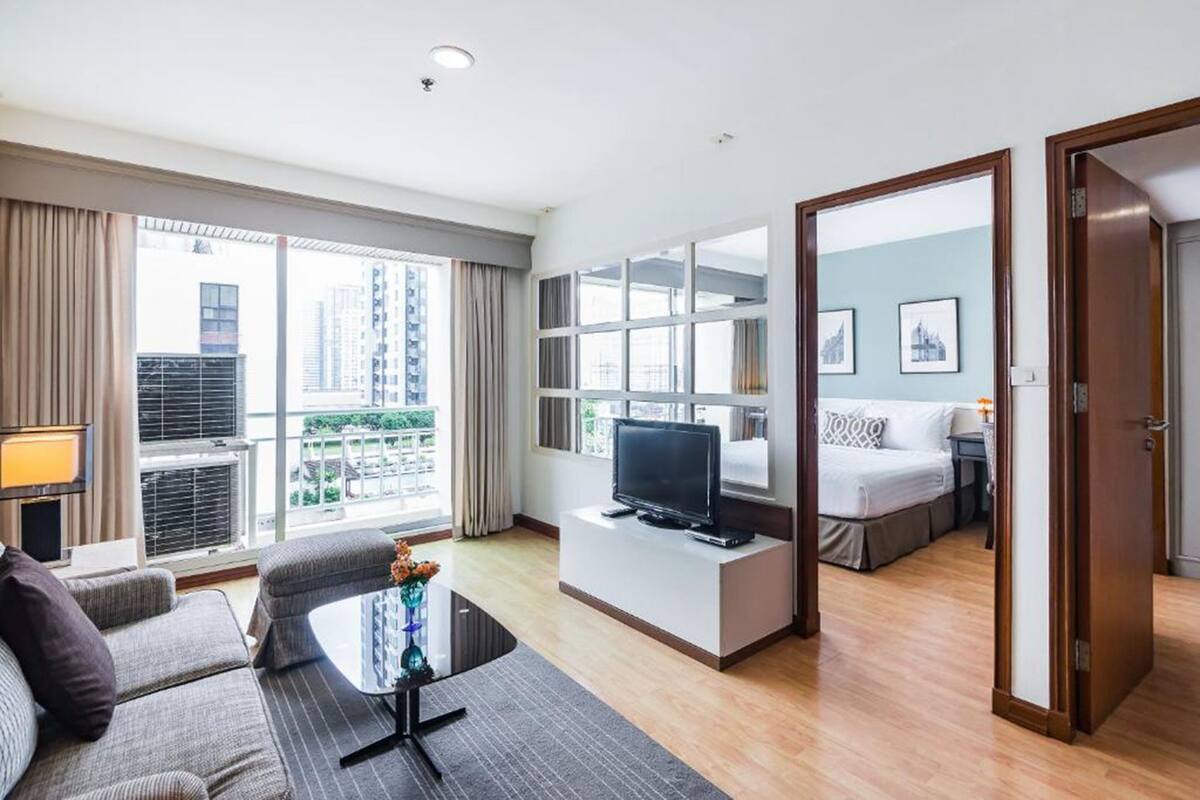A modern hotel room with a sofa, a small table, a TV, a balcony with curtains, and a bed with a nightstand.