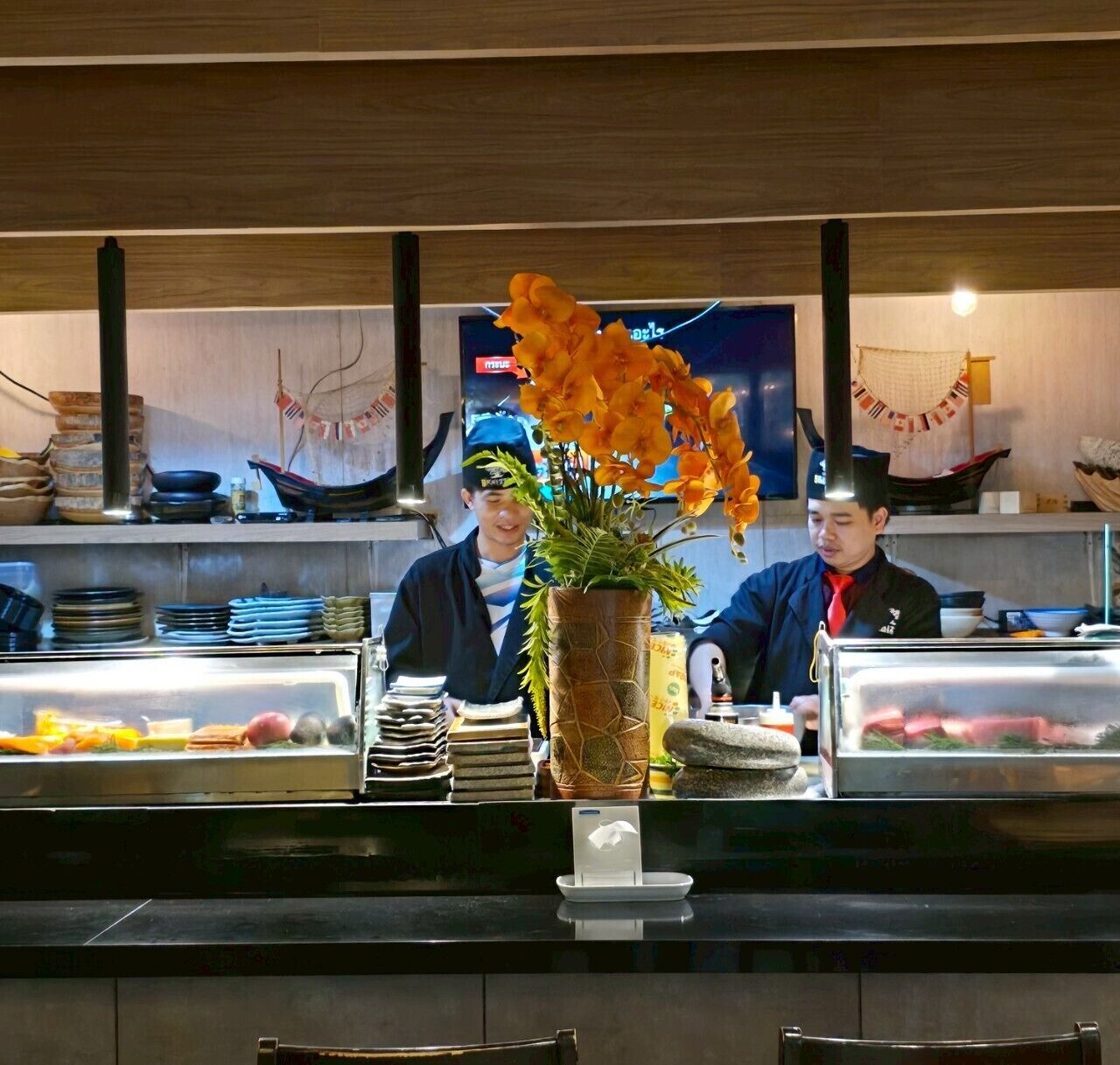 Een sushi restaurant met twee koks, een vitrine met eten, en een decoratieve vaas met bloemen.