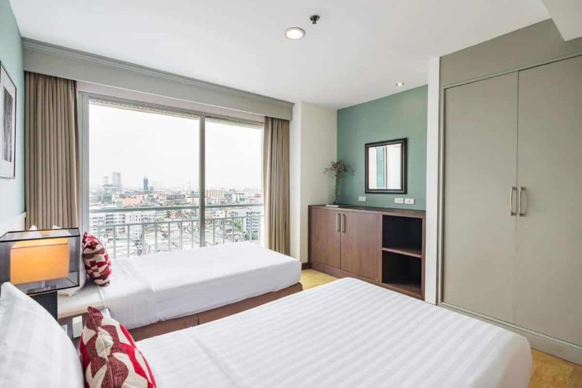 A hotel room with two beds, a large window with a city view, a wardrobe, and a desk with a lamp.