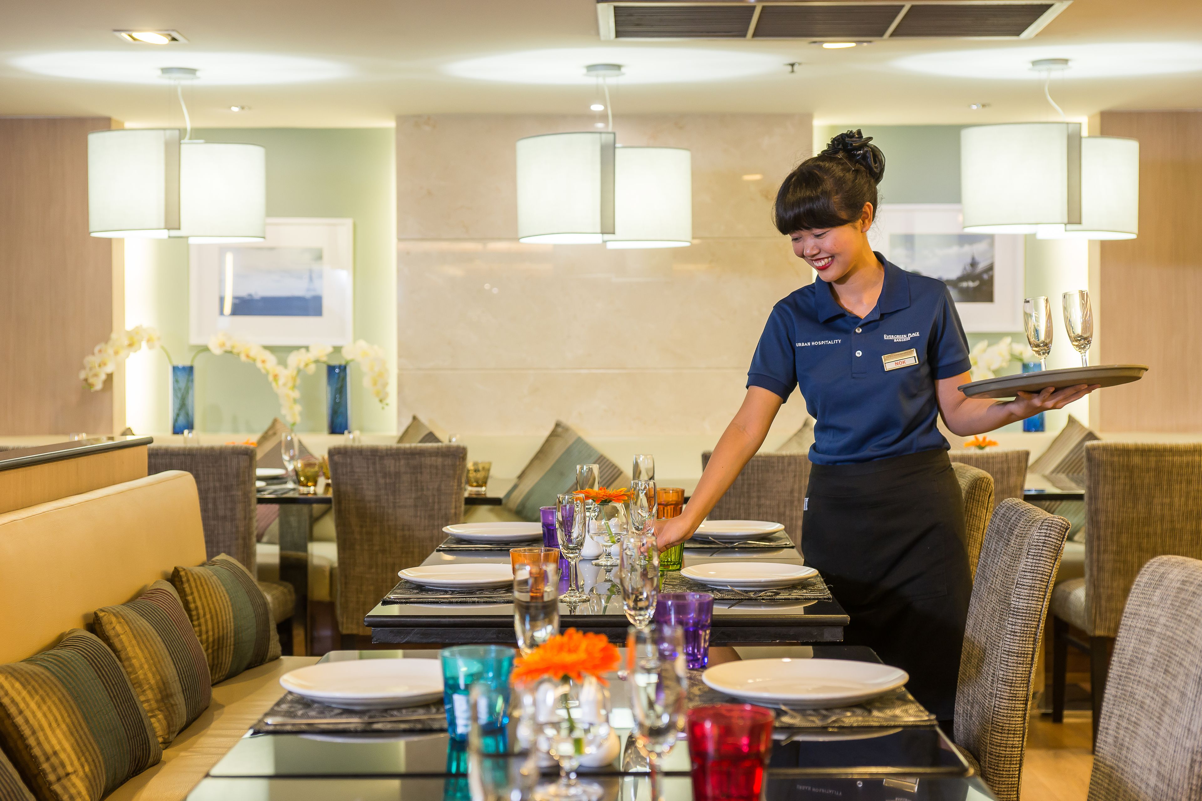 A hotel dining area with tables set for service, a waiter holding a tray, and a modern interior design.