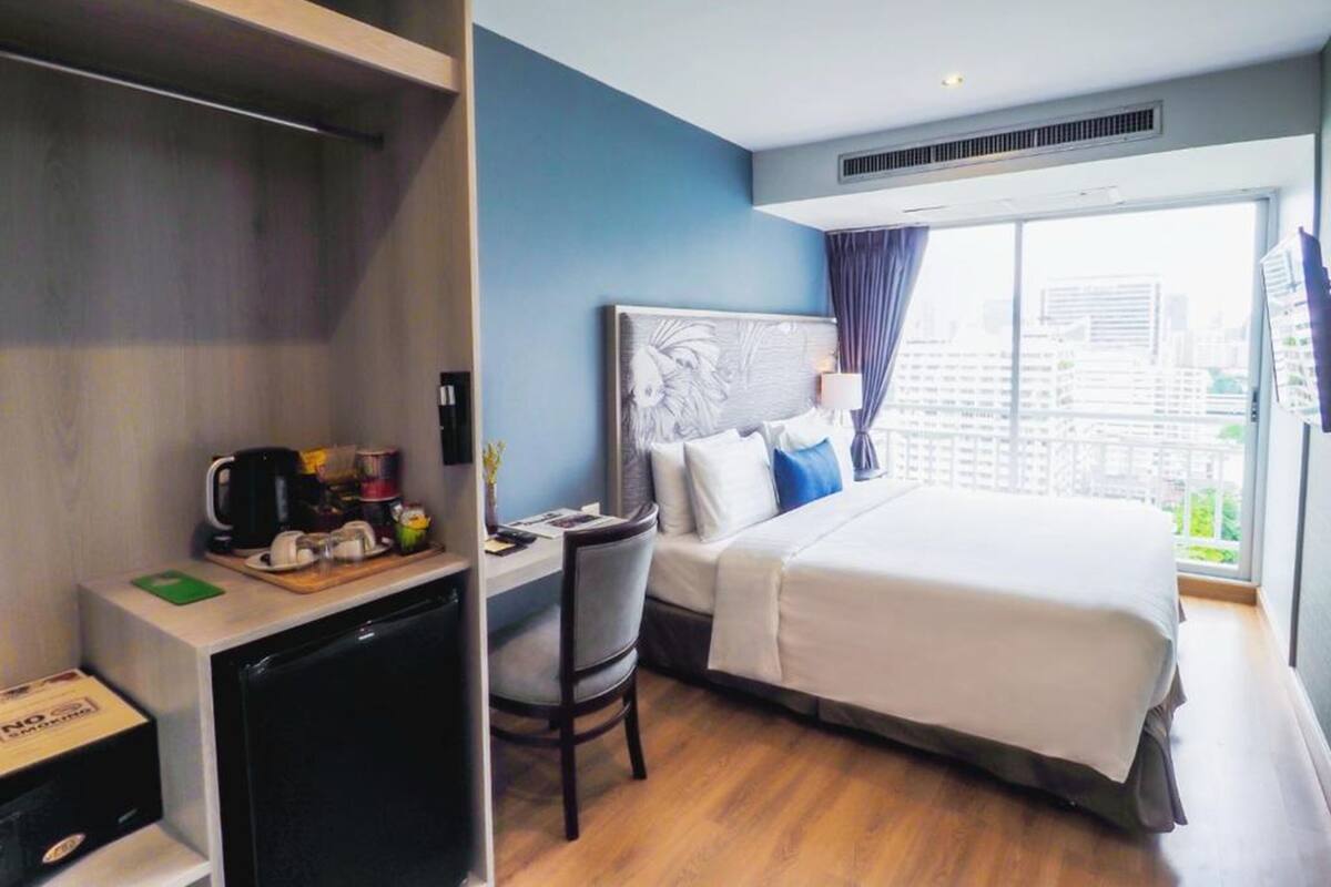 A hotel room with a bed, a desk with a coffee maker, a chair, and a window with a city view.