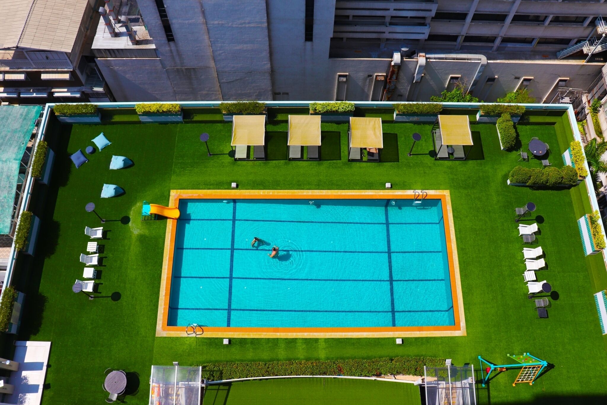 Vista aérea de una piscina en la azotea con un diseño de cuadrícula, rodeada de césped, sillas de descanso y sombrillas.