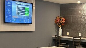 Una sala de conferencias con una pantalla grande mostrando un pronóstico de lluvia, varias mesas y sillas, y un jarrón decorativo con flores en la pared.