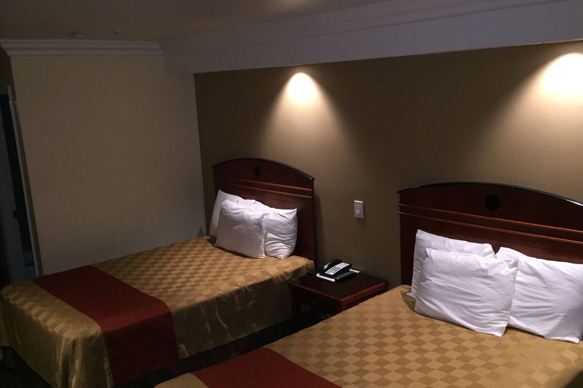 A hotel room with two single beds, each with a wooden headboard and a checkered bedspread. There is a bedside table with a phone and a lamp on one side.