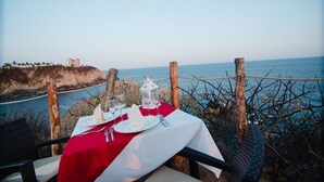 A table set for two with a red and white tablecloth, overlooking a coastal area with a view of the sea and a building on a cliff.
