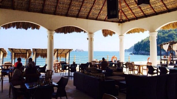 An outdoor dining area with thatched roofs and white columns, overlooking the sea.