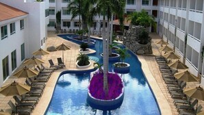 A swimming pool with lounge chairs and umbrellas, surrounded by a building with multiple floors and balconies.