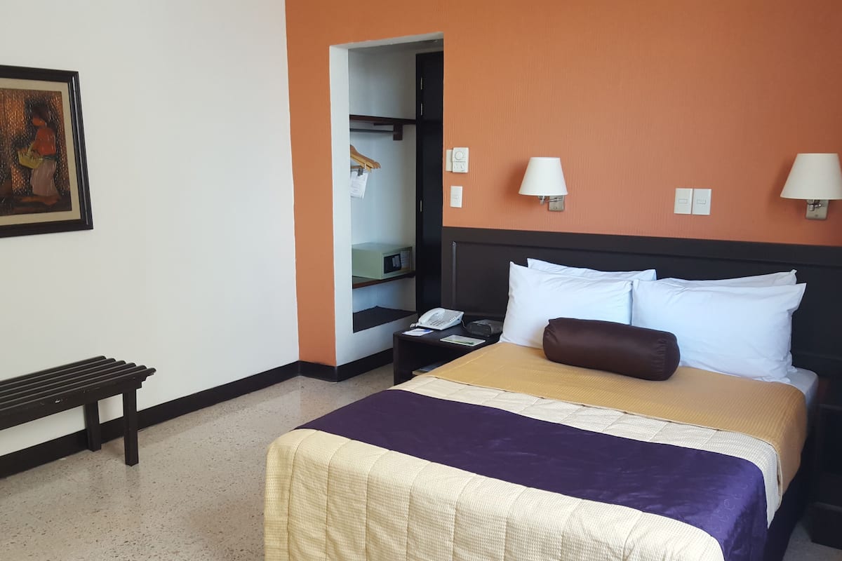 A hotel room with a bed, bedside lamps, a nightstand, a bench, a painting, and a wardrobe.