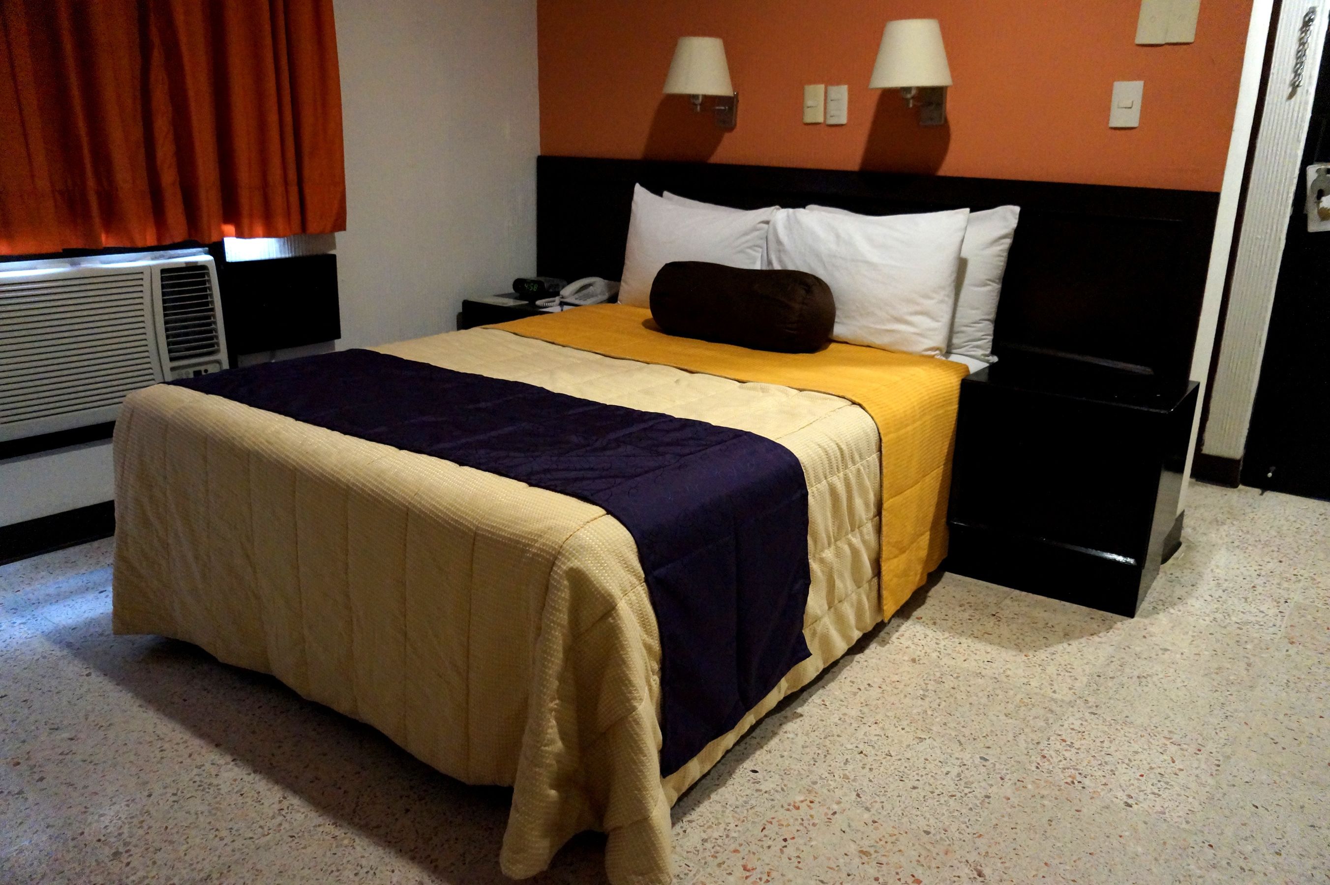 A hotel room with a bed, a nightstand, a lamp, and a wall-mounted heater.