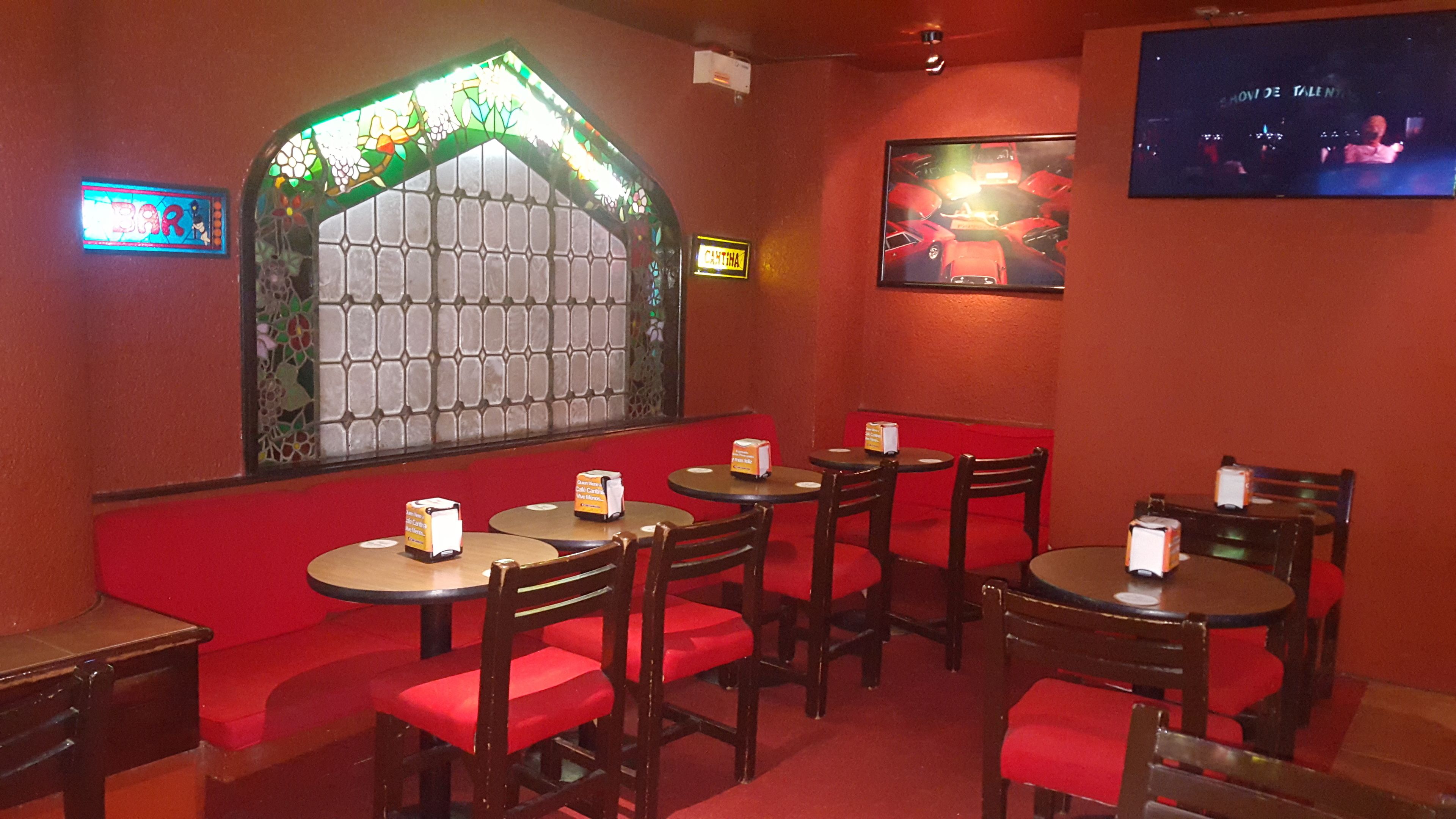 Interior de restaurante con asientos de cuero rojo y mesas redondas, una ventana con vitrales y una pantalla de televisión mostrando una exposición de coches.