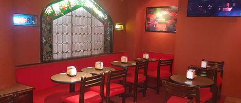 Interior de restaurante con asientos de cuero rojo y mesas redondas, una ventana con vitrales y una pantalla de televisión mostrando una exposición de coches.