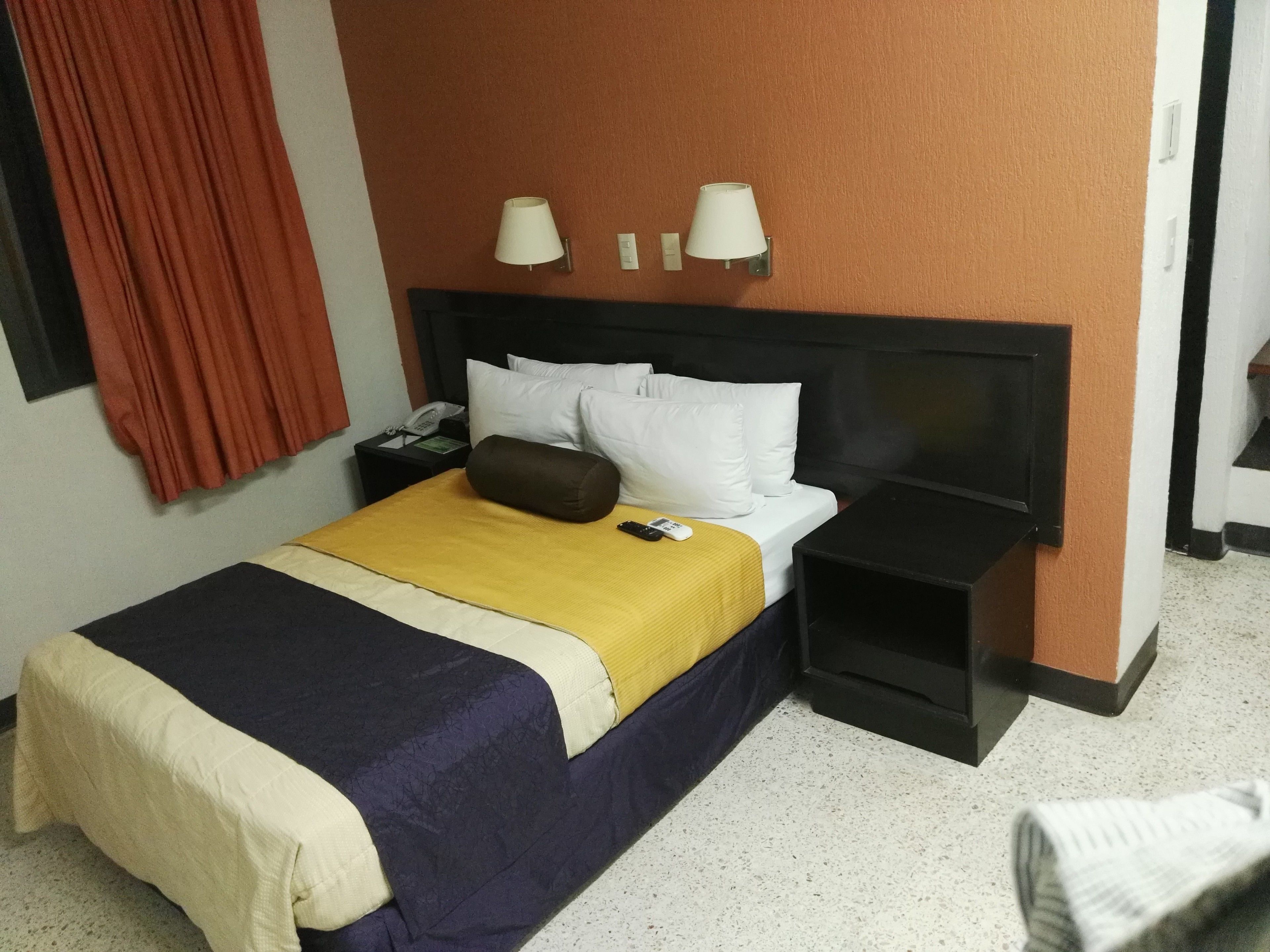 A hotel room with a bed, a nightstand, a lamp, a telephone, and a red curtain.
