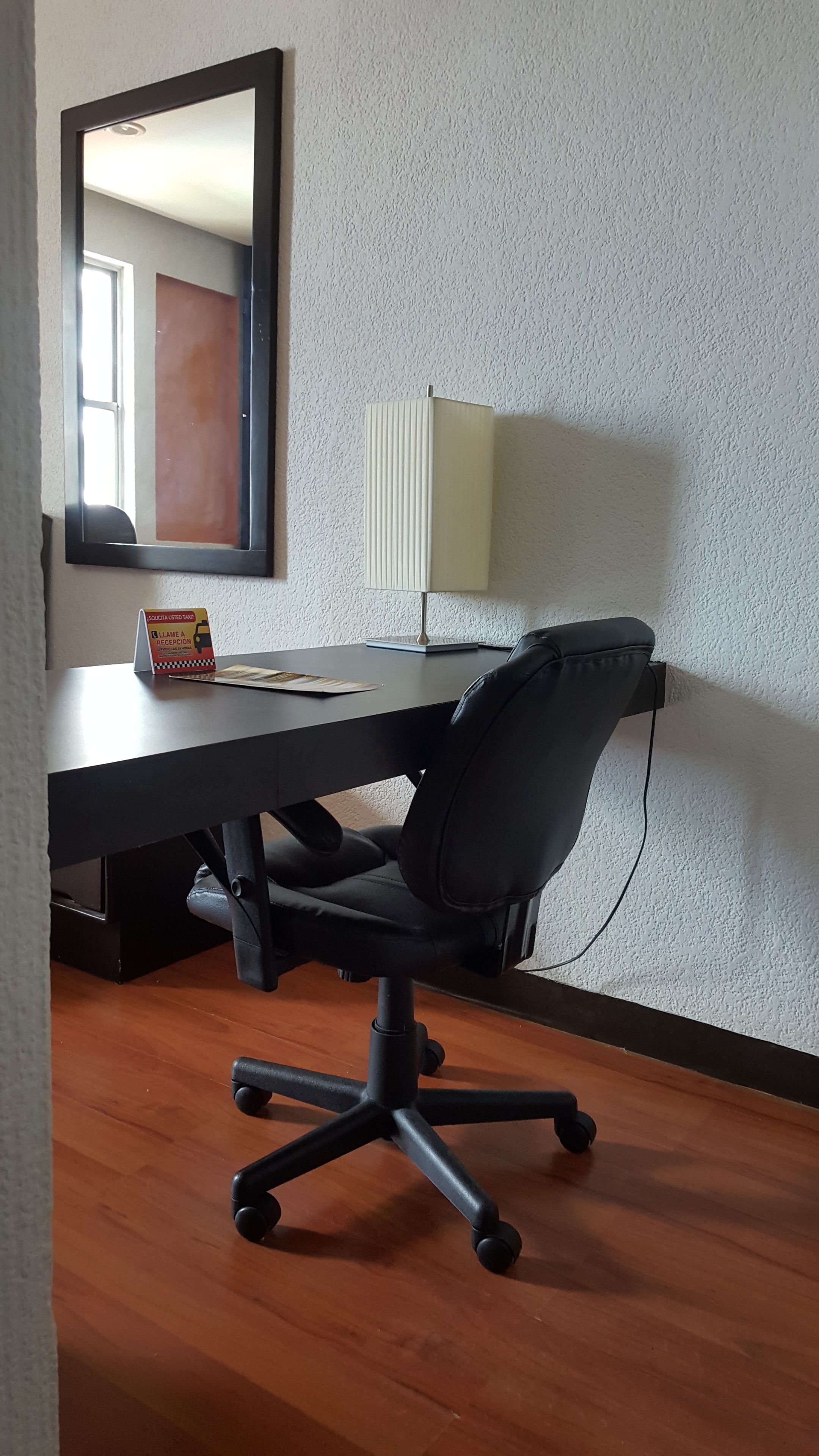Una habitación con un escritorio, una silla de oficina negra, una lámpara y un espejo en la pared.
