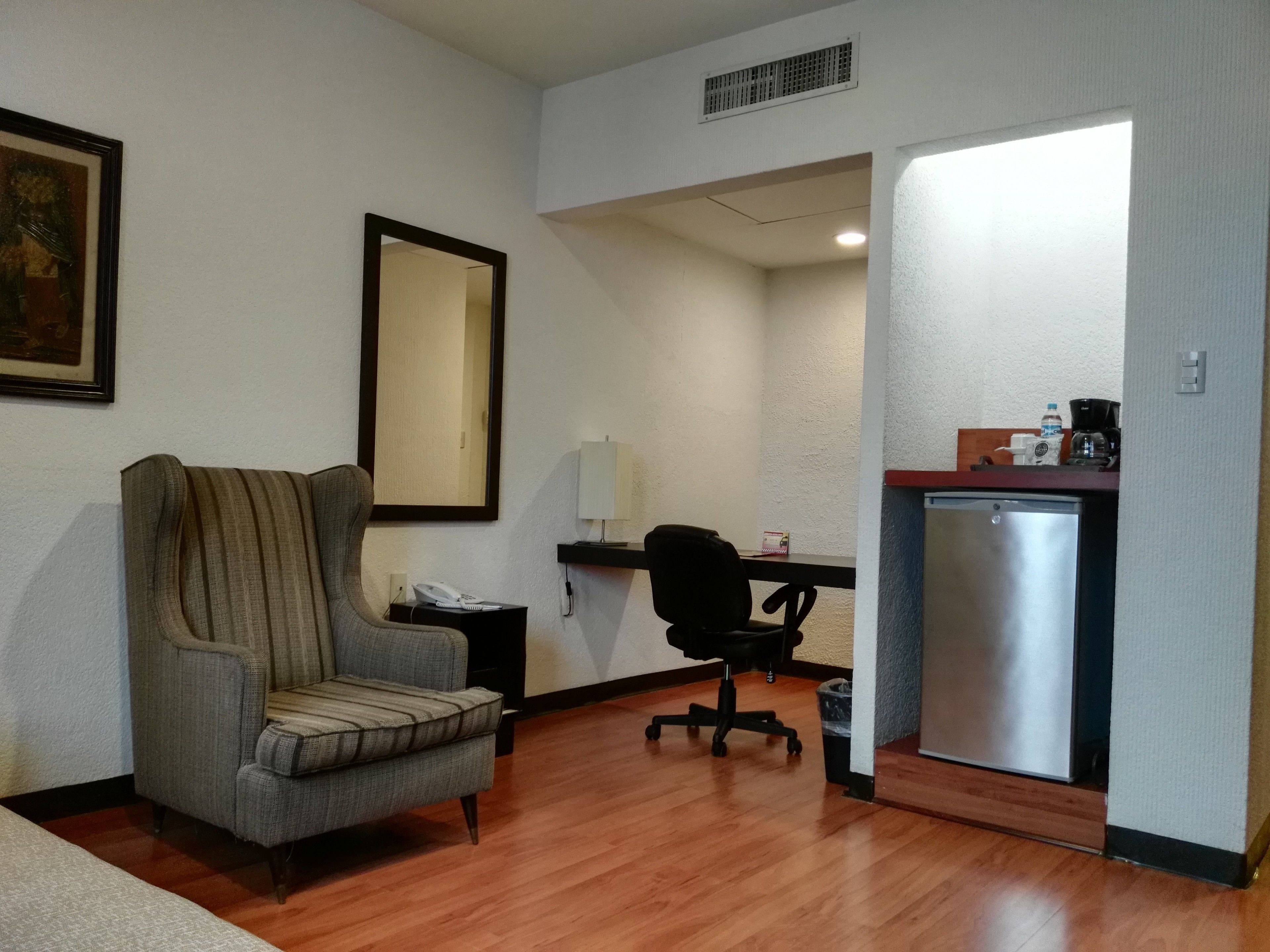 A hotel room with a grey armchair, a desk with a black office chair, a small refrigerator, and a painting on the wall.