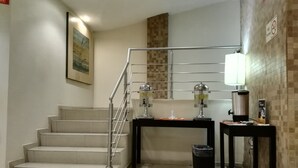 Uma escadaria com corrimão de metal, um cesto de lixo, uma mesa com uma máquina de café e uma placa fixada na parede.