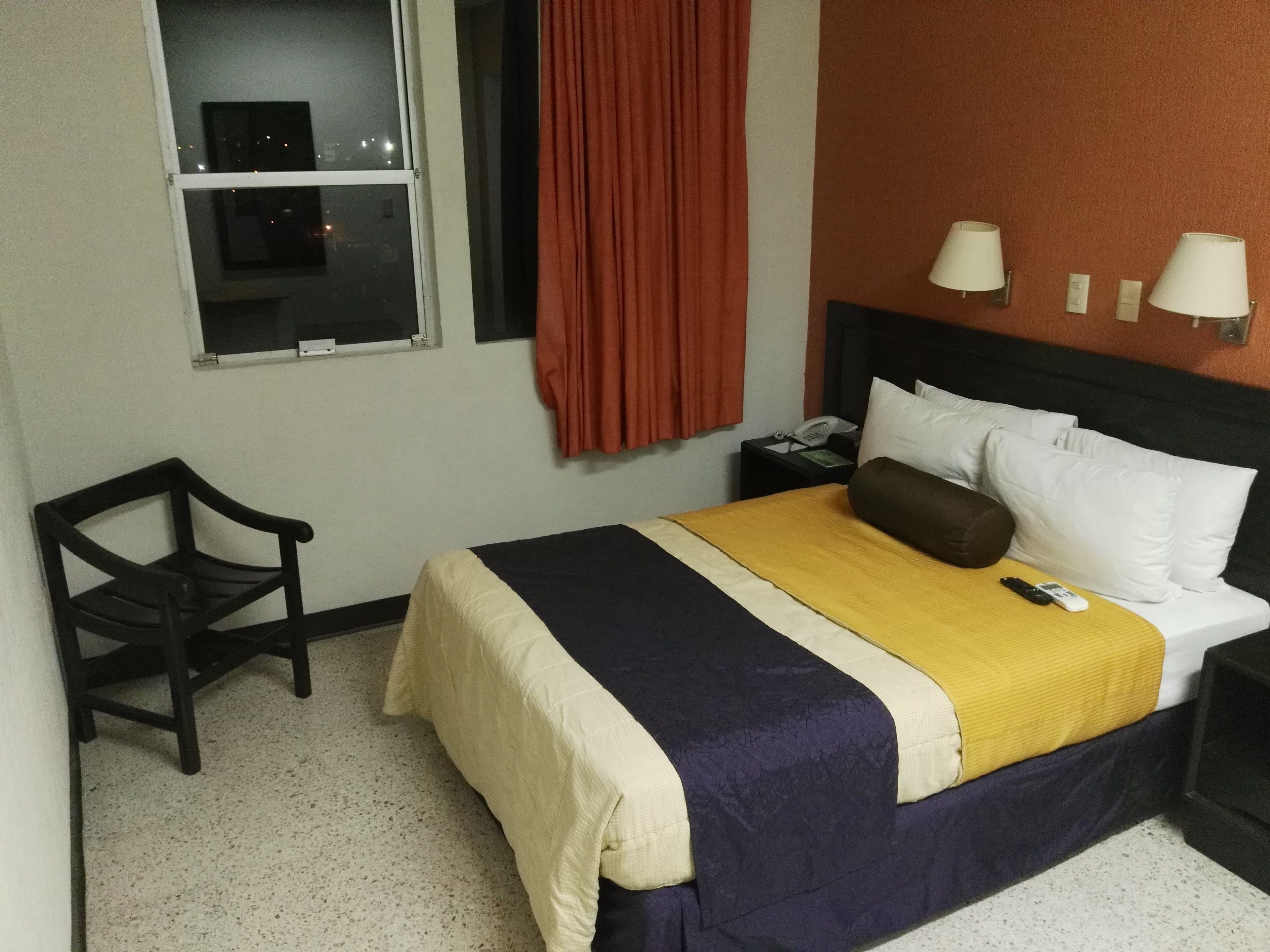 Habitación de hotel con cama, mesita de noche, silla, ventana con cortinas y dos lámparas de pared.