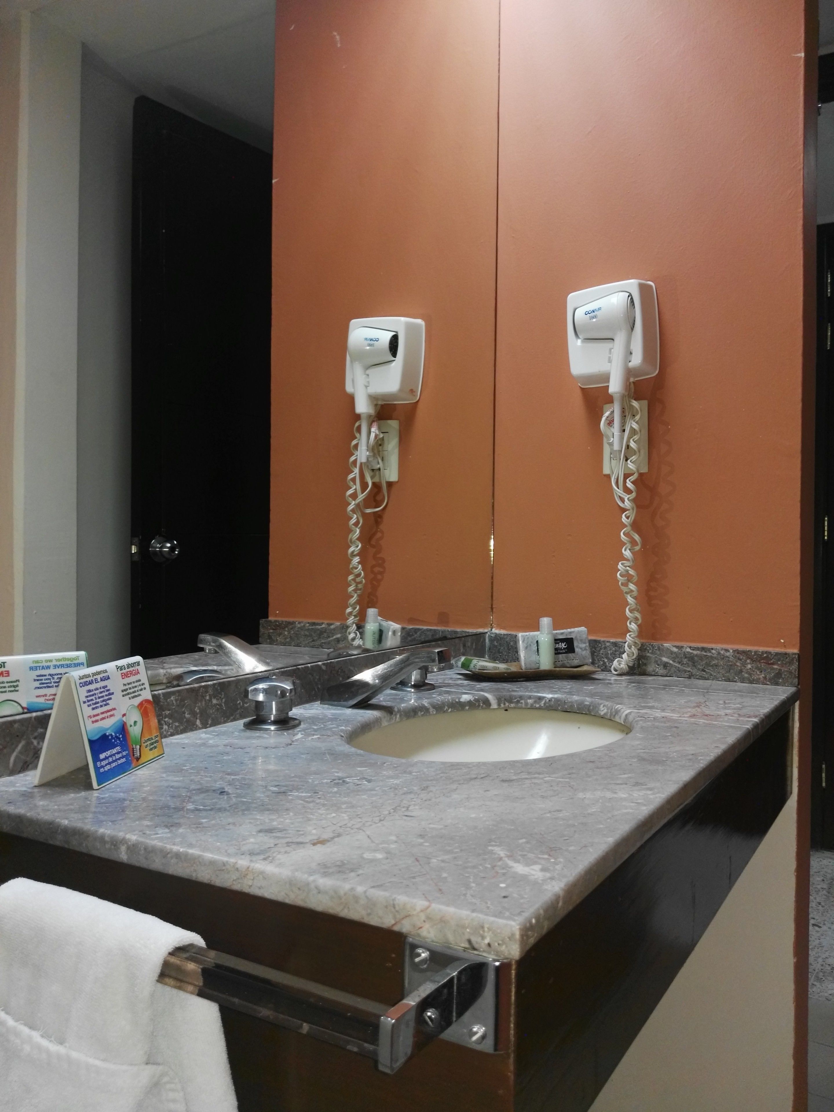 Une salle de bain équipée d’un lavabo, de deux sèche-cheveux fixés au mur, d’un porte-serviettes et d’un distributeur de savon liquide.
