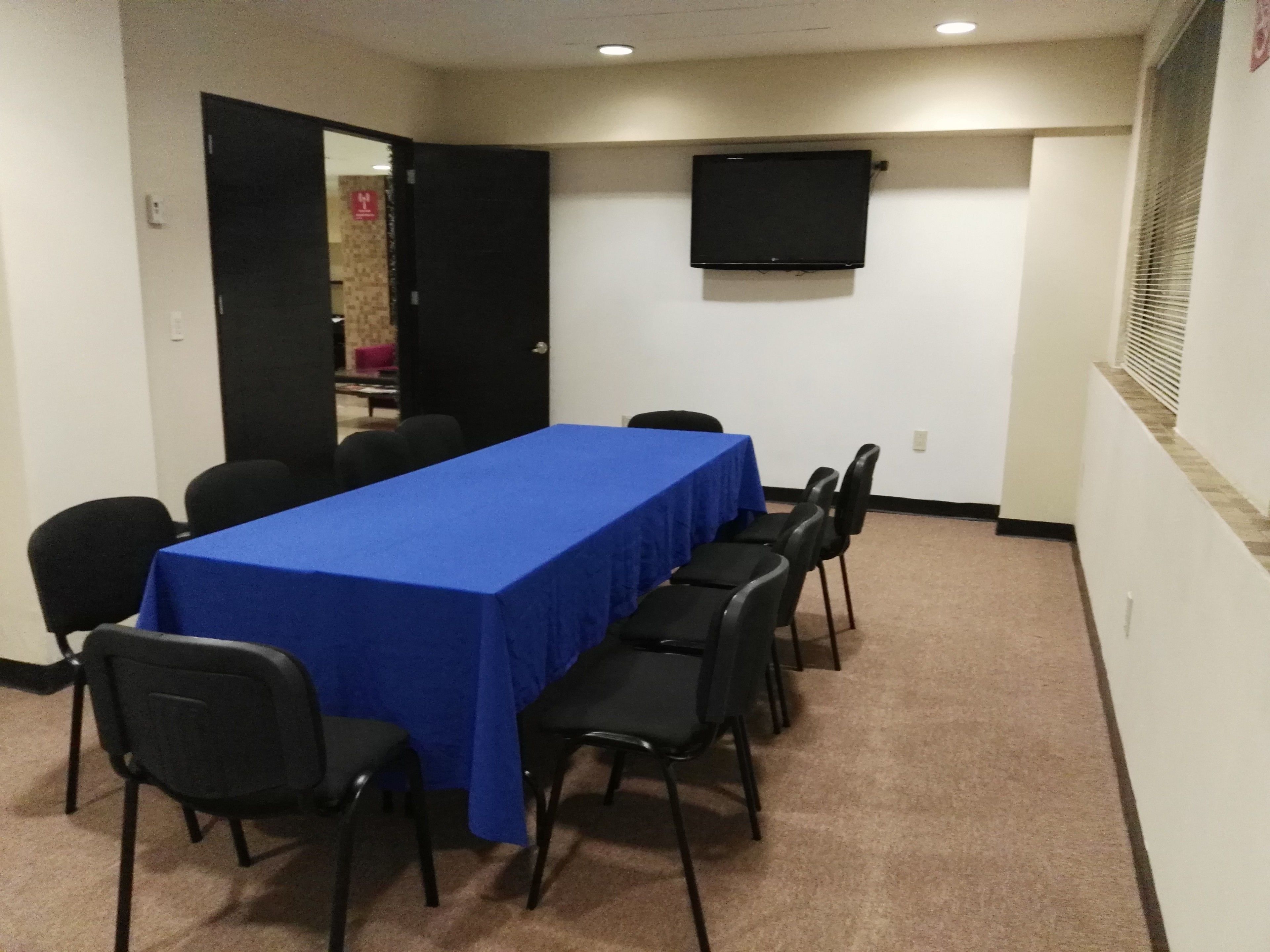 A conference room with a long table covered in blue, surrounded by black chairs, a flat-screen TV on the wall, and a door leading to another room.