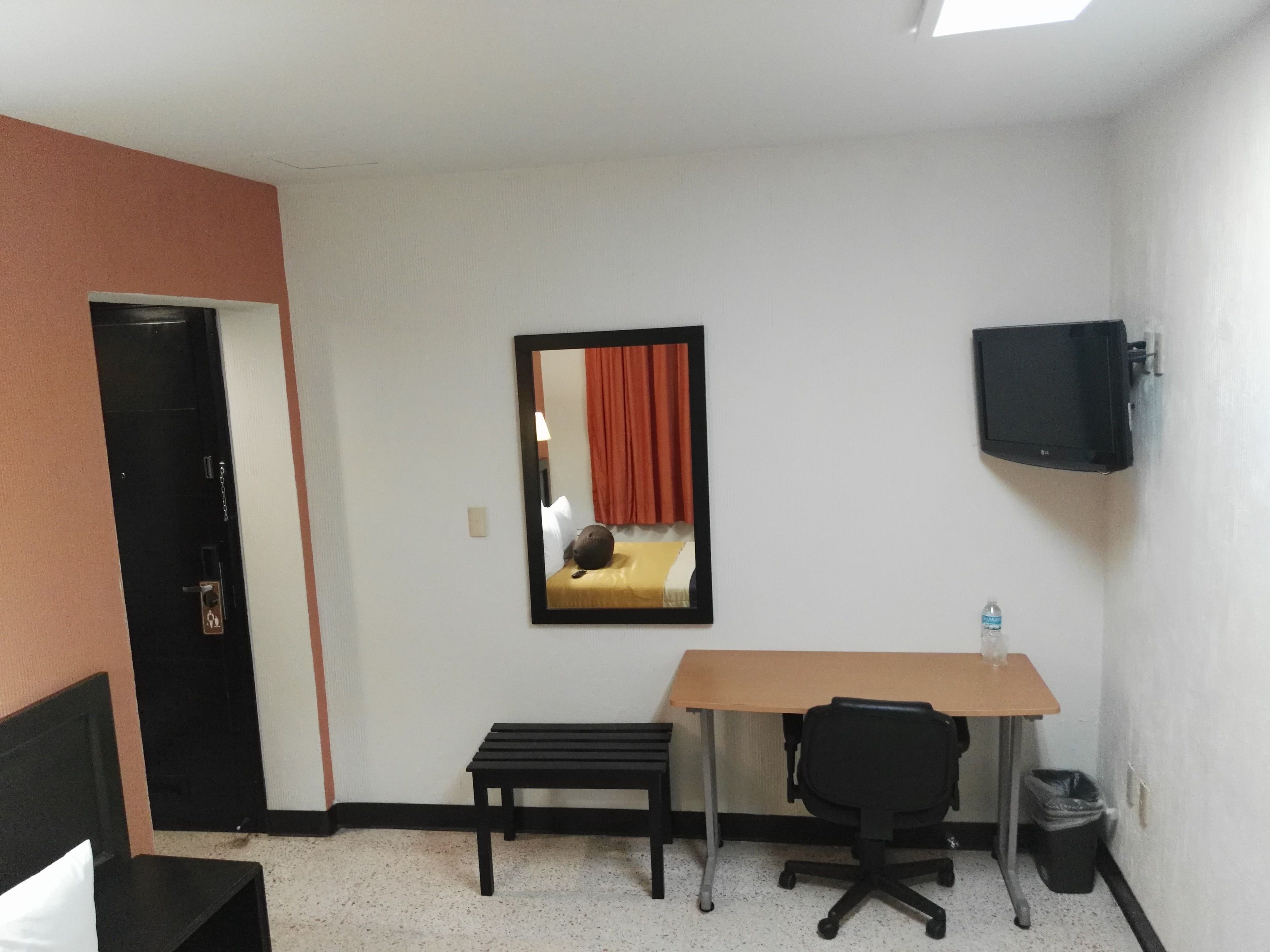A hotel room with a bed, a desk with a chair, a TV mounted on the wall, and a trash bin.