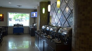 Uma área de buffet com diversas estações de autoatendimento, uma tela de televisão e um balcão coberto por uma toalha azul.