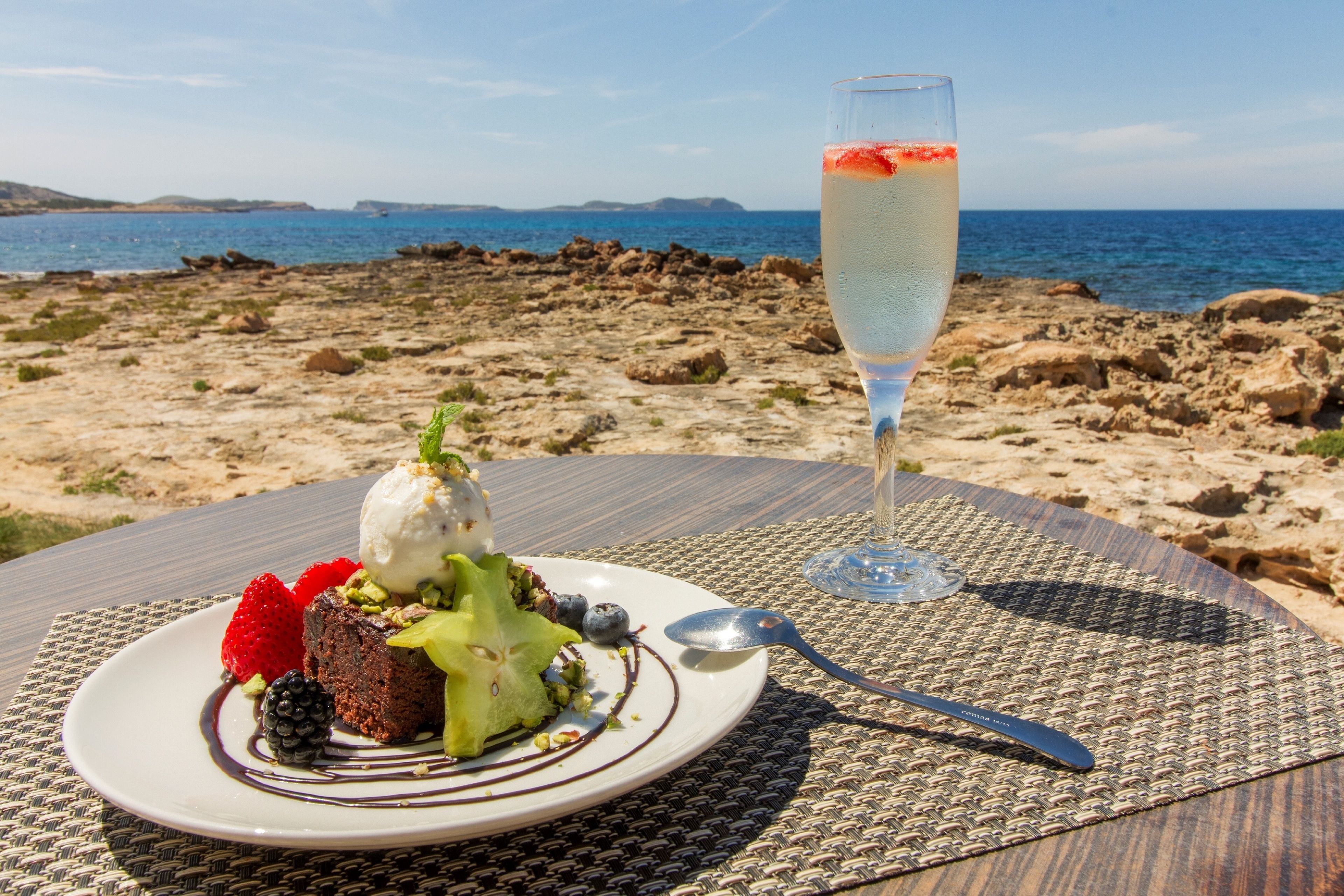 A dessert plate with ice cream, fruits, and nuts on a table with a view of the sea and rocky landscape.