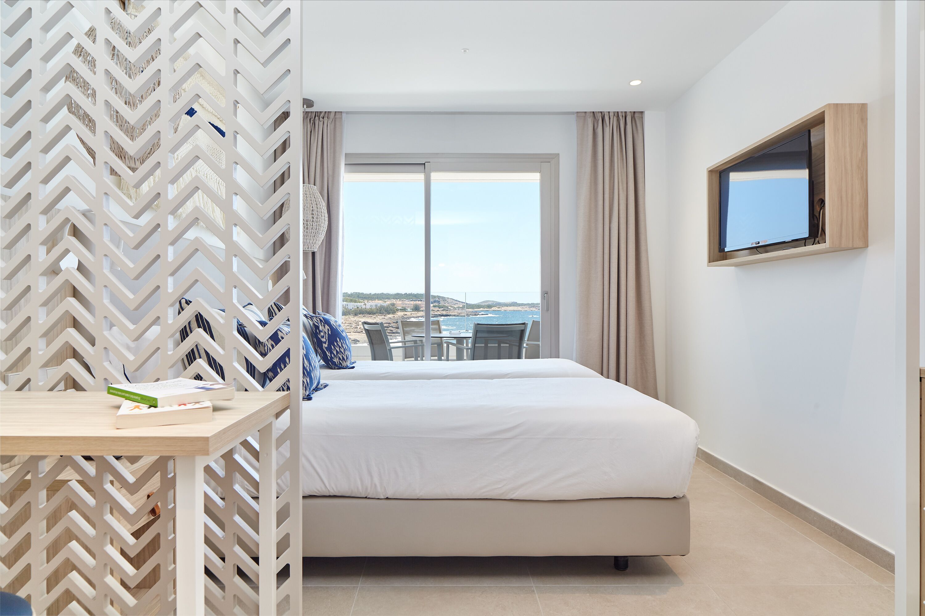 A hotel room with a bed, a TV mounted on the wall, a view of the sea through a sliding door, and a table with a book.